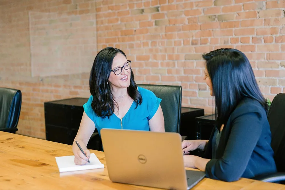 Two women seated at a wooden table in an office having a conversation, one writing in a notebook and the other using a laptop.