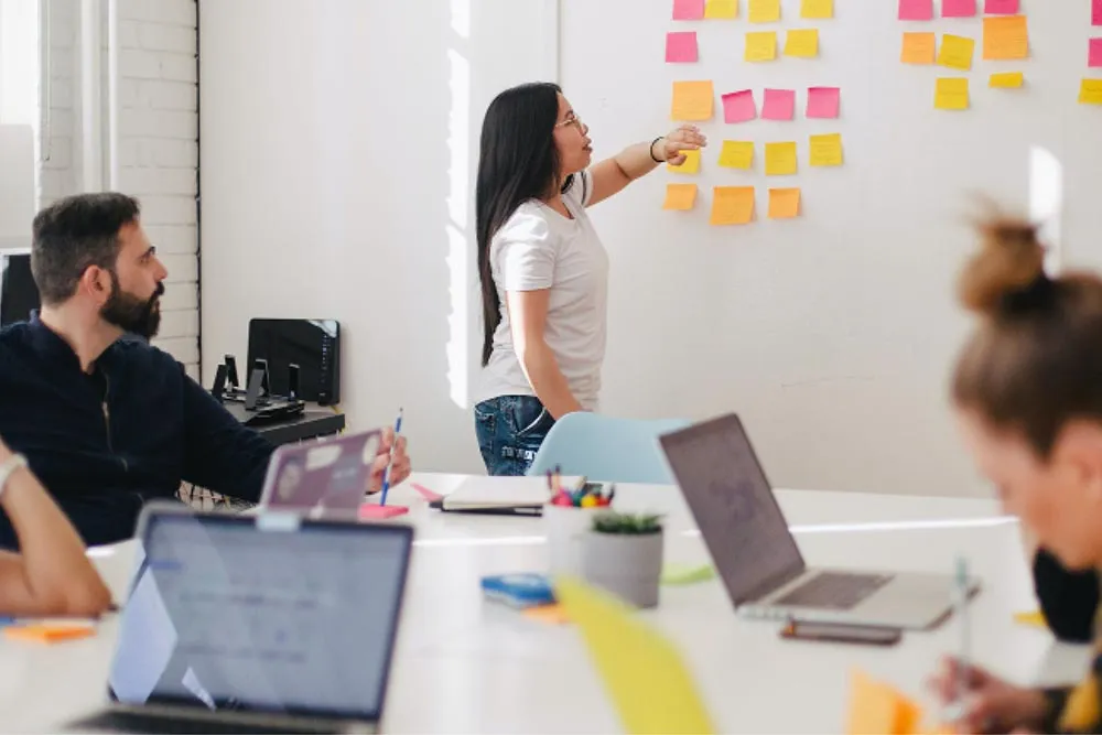 A woman points to colorful sticky notes on a whiteboard while colleagues listen and take notes in a meeting room.
