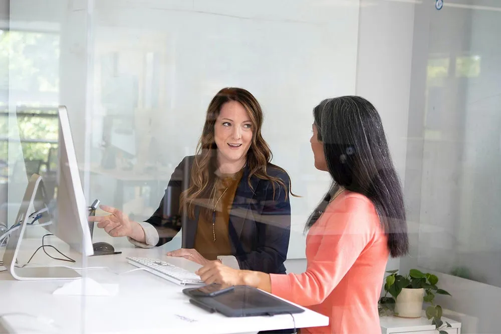 Two women having a discussion in a modern office, one pointing at a computer screen.