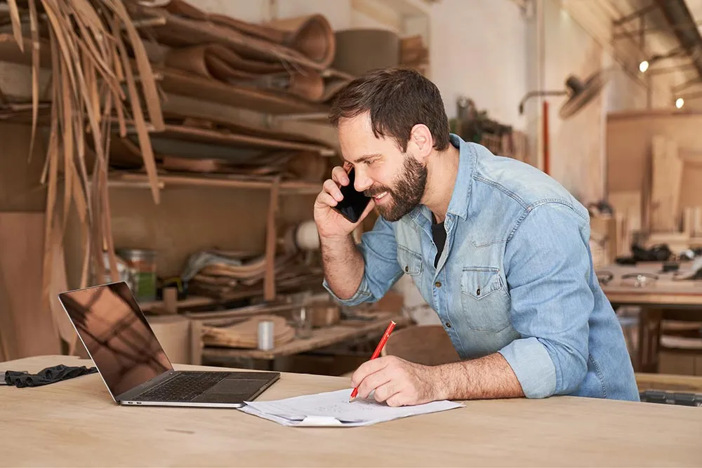 Man in a denim shirt talking on a smartphone while writing on papers at a wooden workbench in a workshop.