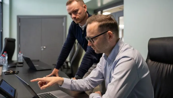 Two men in an office, one sitting and pointing at a laptop screen while the other stands and watches attentively.