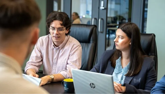 Two professionals sitting at a conference table engaged in discussion, with a laptop and coffee mug in front of them.