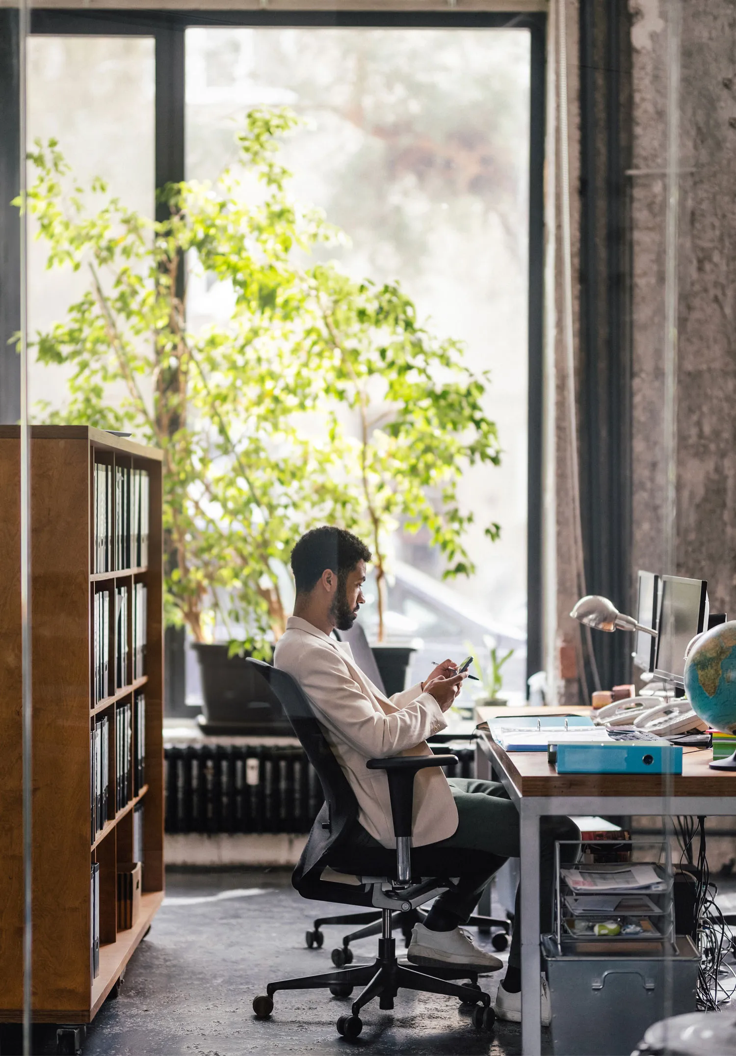 Man in beige blazer sitting on office chair and using smartphone at desk with computer monitors, globe, and office supplies.