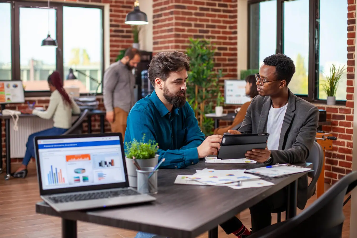 Two professionals sitting at a conference table engaged in discussion, with a laptop and coffee mug in front of them.