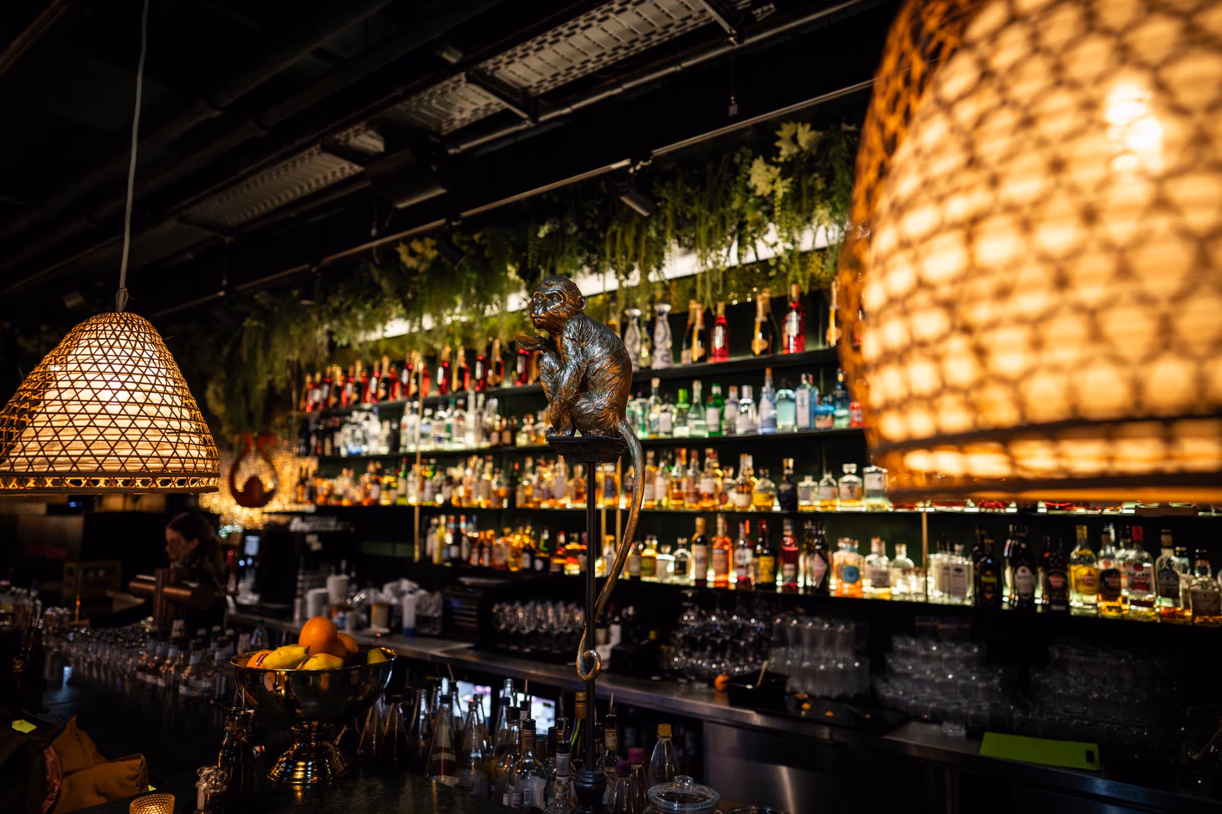 Bar mit einem Regal voller bunter Spirituosenflaschen, hängenden Korblampen und einer dekorativen Affenskulptur auf einem Ständer.