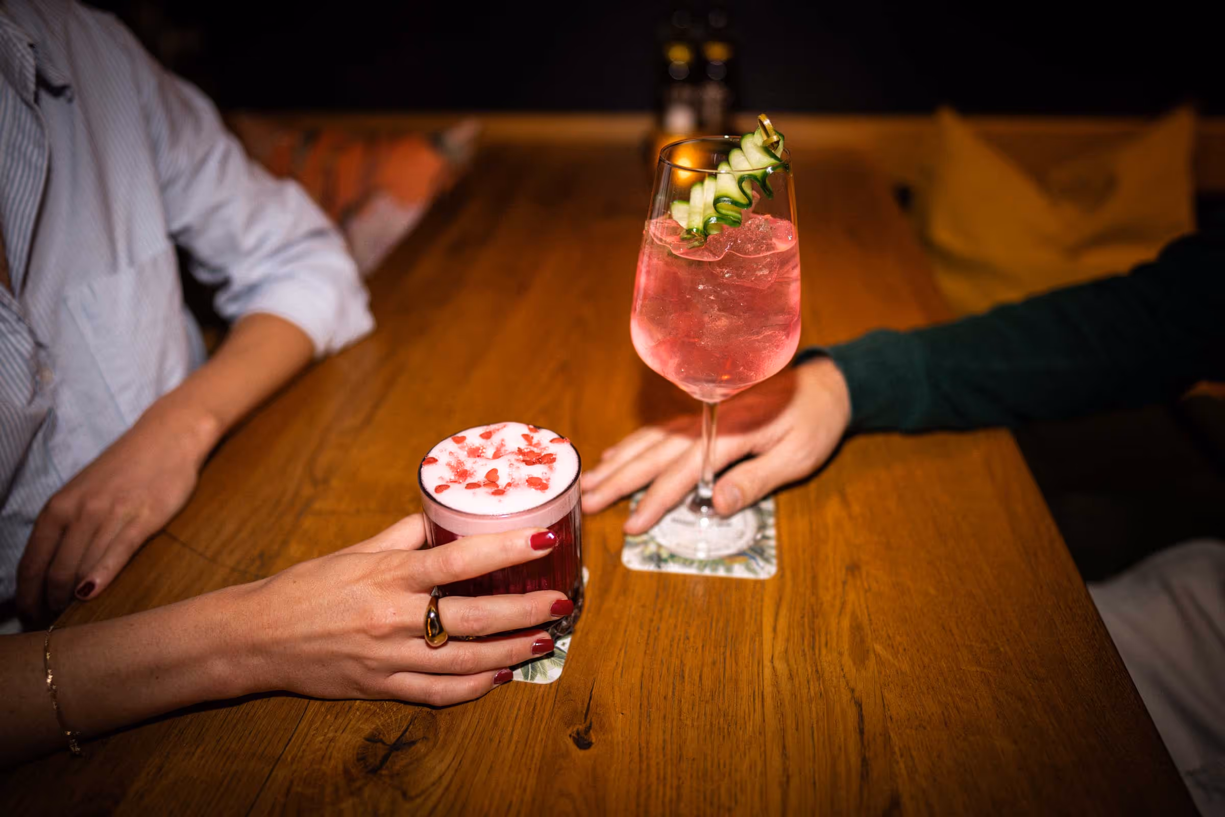 Zwei Personen sitzen an einem Holztisch mit jeweils einem pinkfarbenen Cocktail, einer im Tumbler mit Schaum und Beeren, der andere im Weinglas mit Gurkengarnitur.