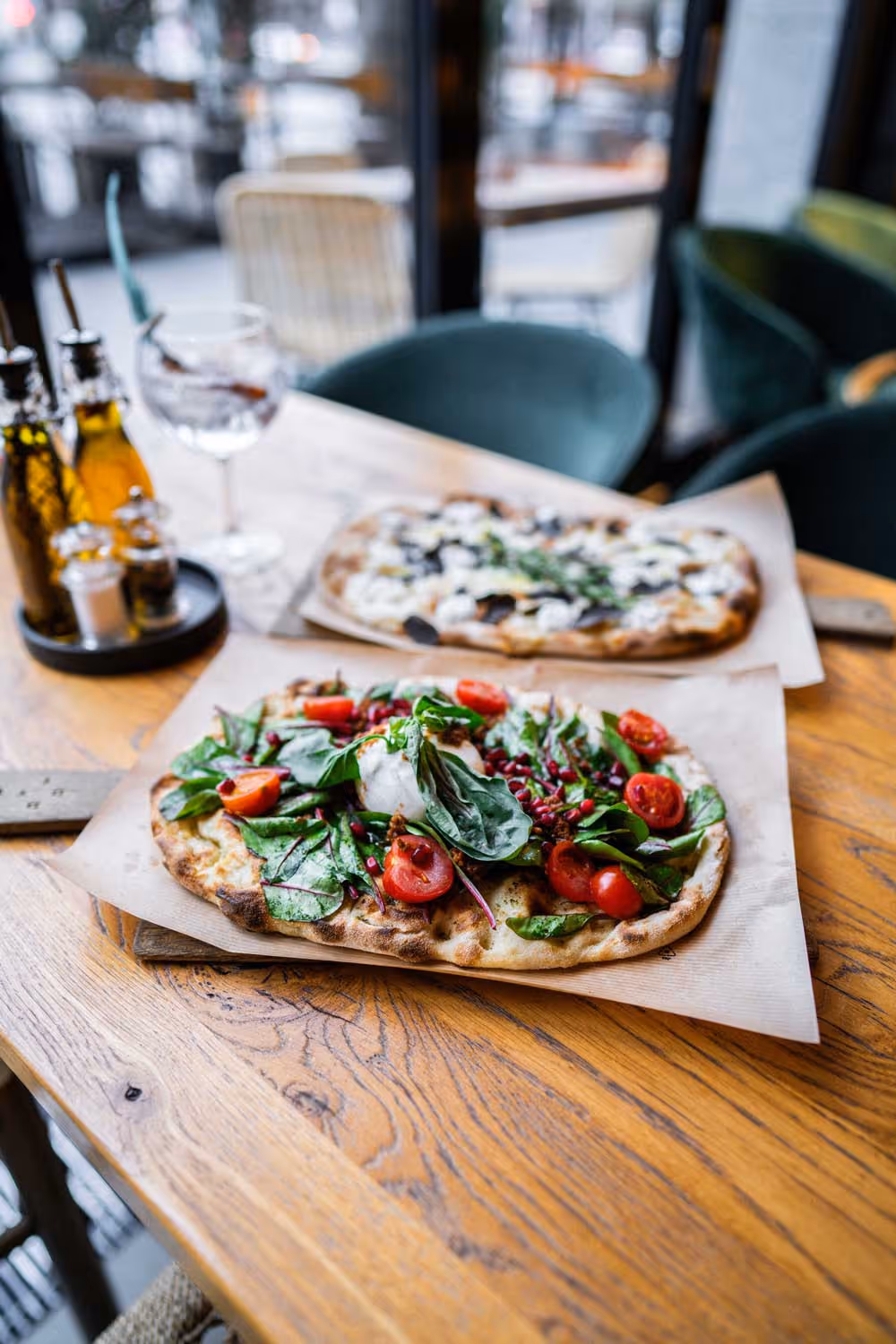 Zwei Flammkuchen auf Holztisch, einer belegt mit Spinat, Cherrytomaten und Pomegranatekernen, im Hintergrund Gewürzflaschen und ein Getränk in einem Glas.