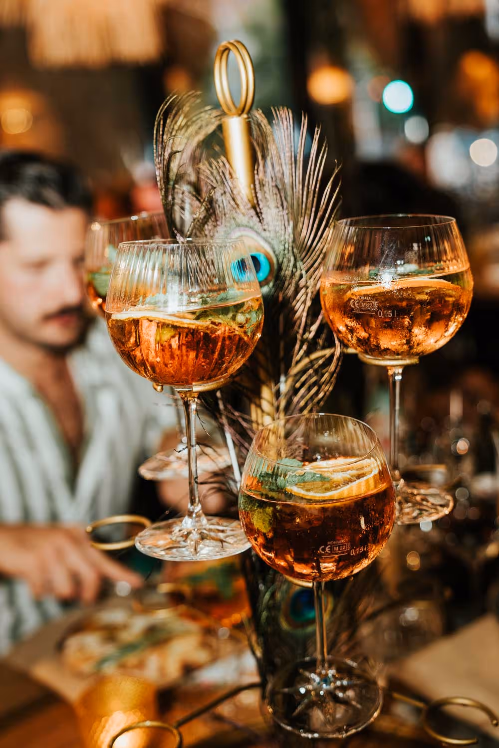 Drei mit Orangenscheiben und Minze dekorierte Aperol-Gläser auf einem Ständer mit Pfauenfedern vor unscharfem Hintergrund mit einer Person.