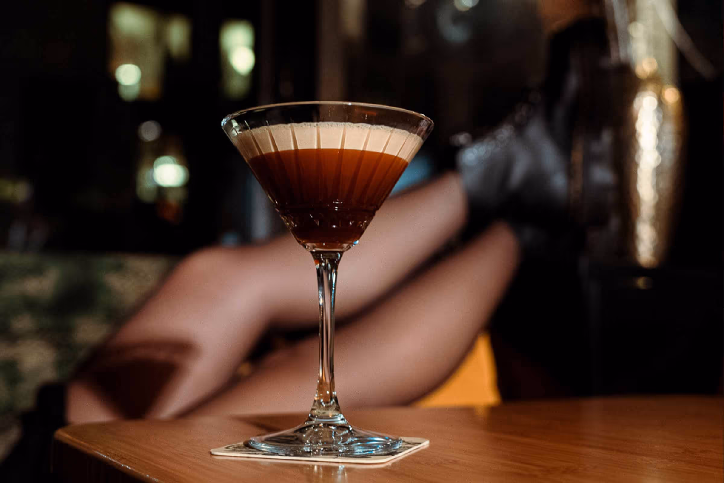 Cocktailglas mit braunem Getränk auf Holzoberfläche, im Hintergrund unscharfe Beine mit schwarzen Stiefeln.