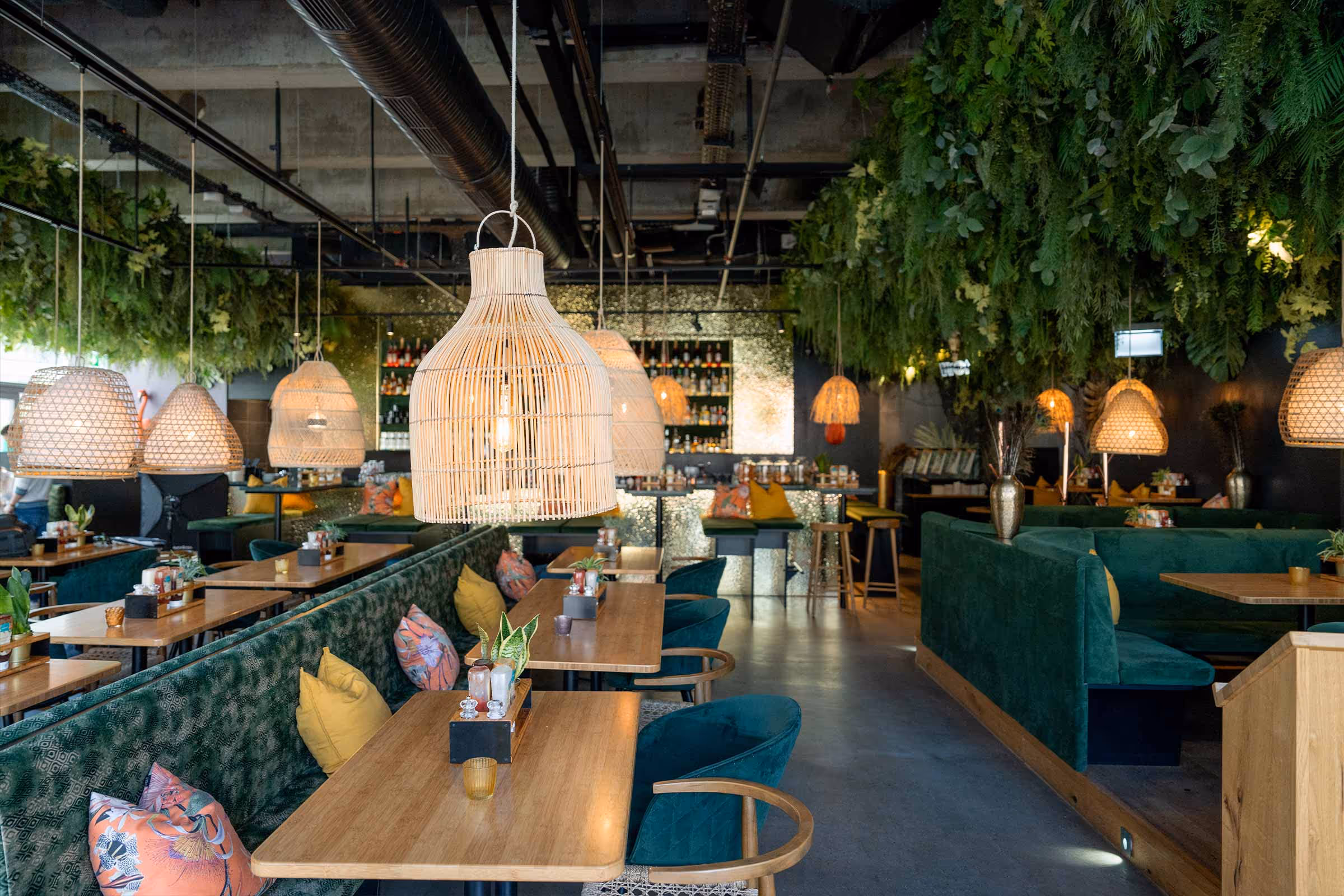Modernes Restaurant mit grünen Samt-Sitzbänken, Holztischen, dekorativen Kissen und hängenden Lampen aus Korbgeflecht unter einer Decke mit grünen Pflanzen.