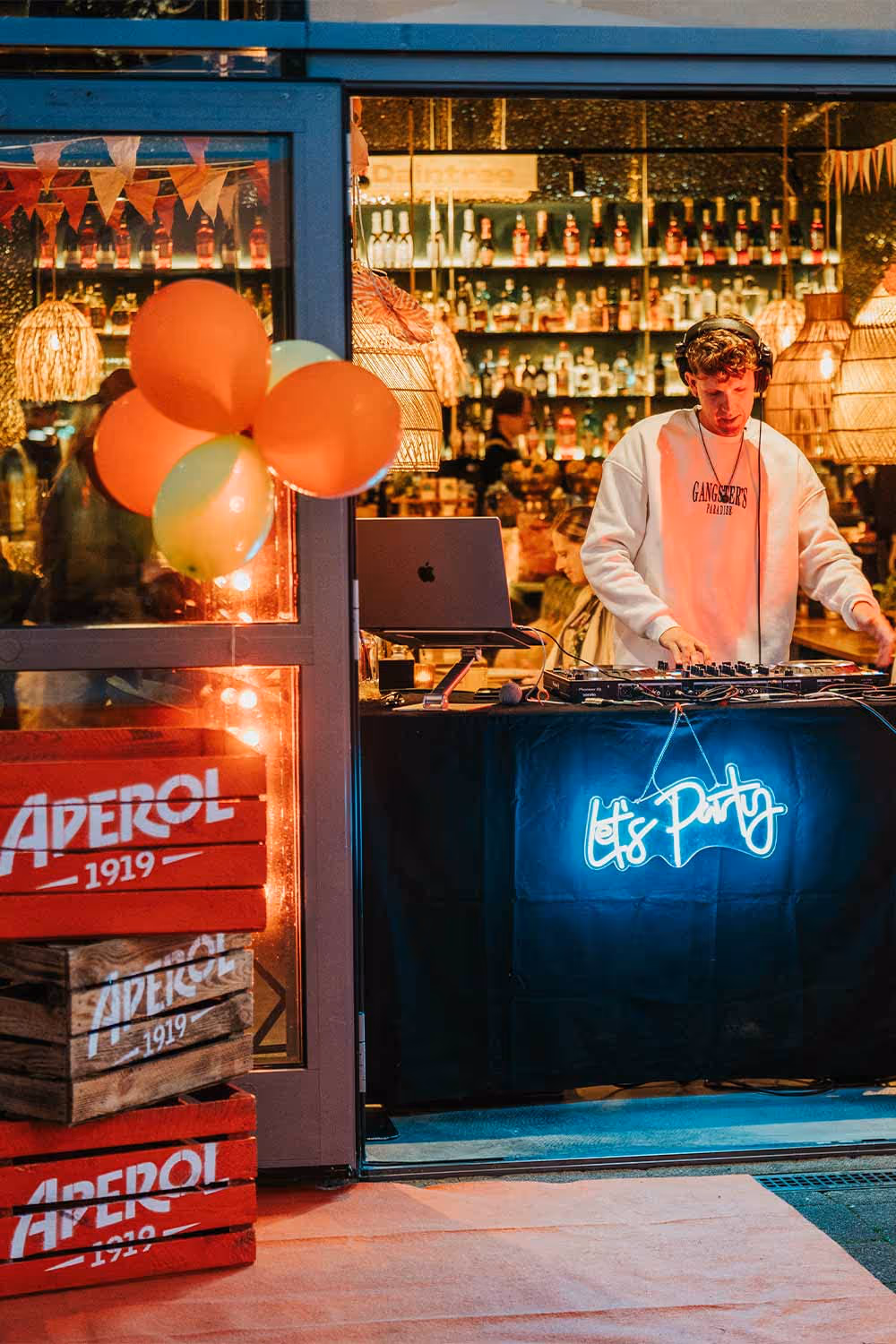 DJ mit Kopfhörern steht an einem Mischpult in einem festlich beleuchteten Raum mit Aperol-Kisten und bunten Luftballons.