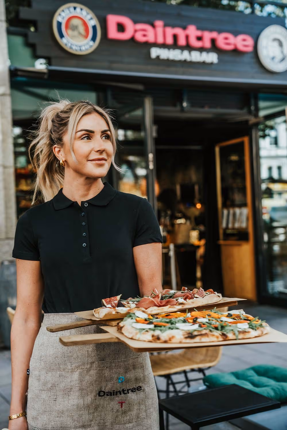 Kellnerin trägt zwei Holztabletts mit frisch belegten Pizzas vor einem Restaurant namens Daintree.