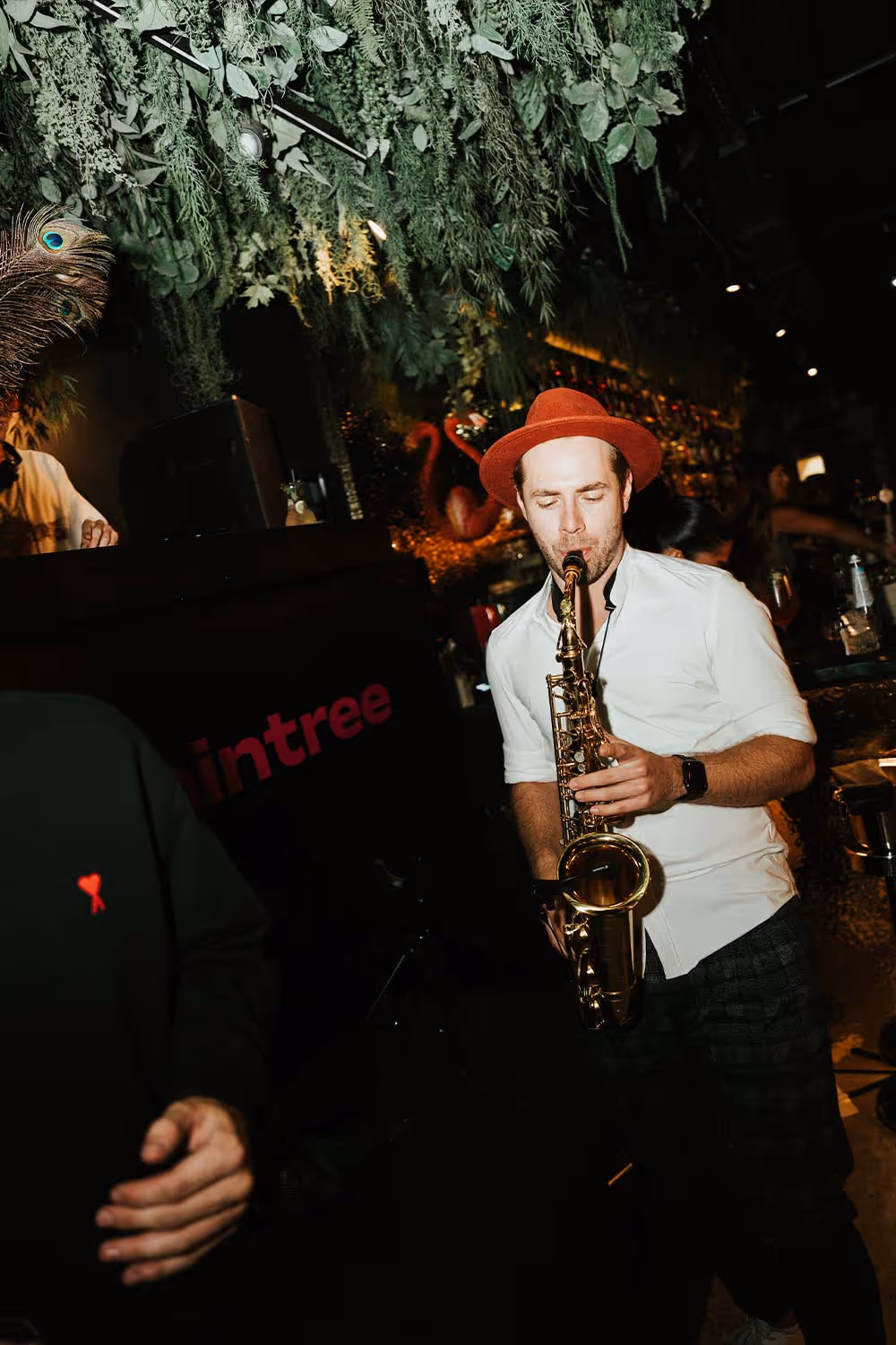 Mann in weißem Hemd und rotem Hut spielt Saxophon in einer Bar mit hängenden grünen Pflanzen.