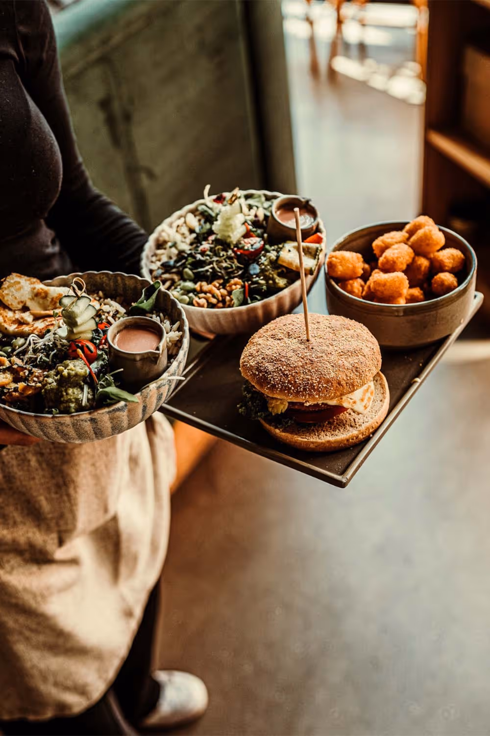 Person trägt Tablett mit Burger, Schüssel mit frittierten Snacks und zwei Schalen mit gemischtem Salat und Dips.