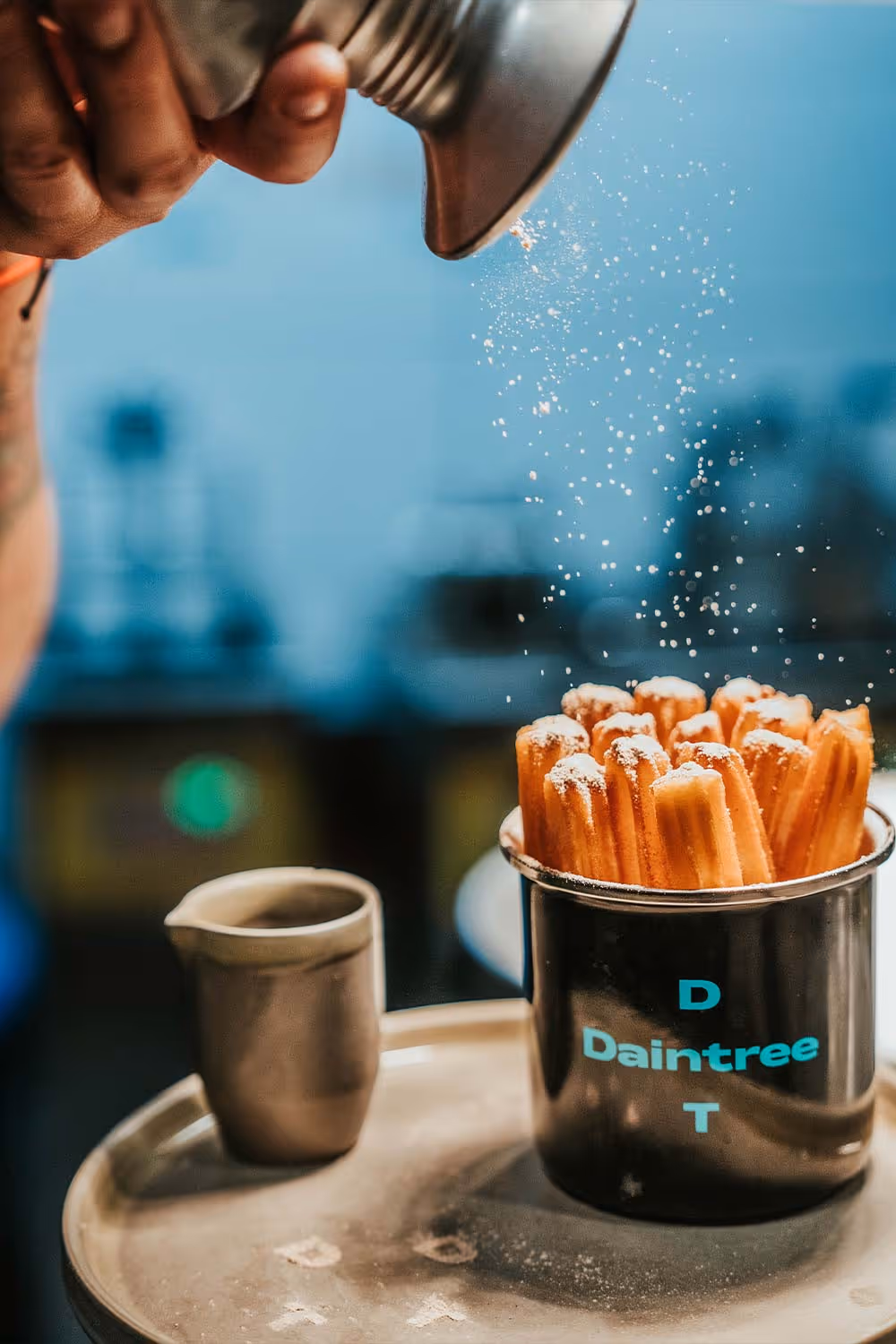 Hand bestreut Puderzucker über einem Becher mit Churros neben einem kleinen Metallbecher auf einem Tablett.