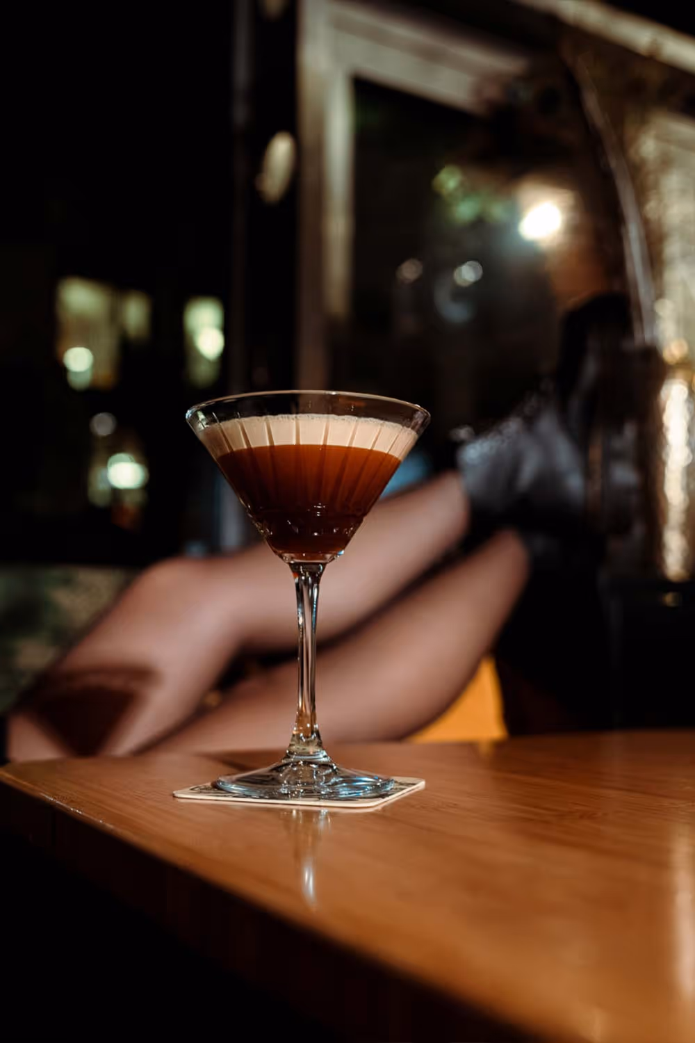 Cocktailglas mit braunem Getränk auf einem Holztisch, unscharfe Beine einer Person im Hintergrund.