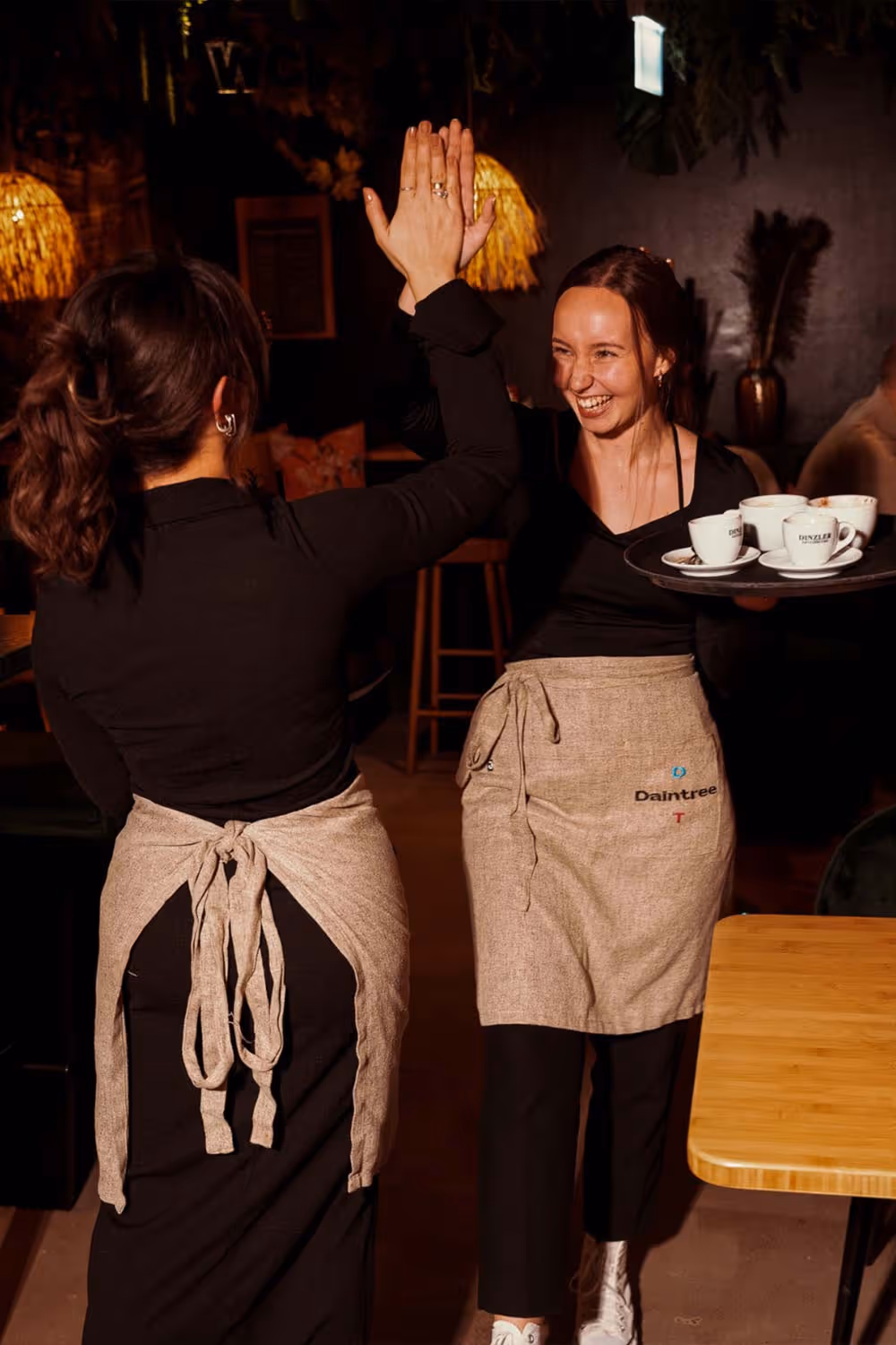 Zwei Kellnerinnen in einem Restaurant klatschen sich lächelnd ab, eine trägt ein Tablett mit Kaffeetassen.