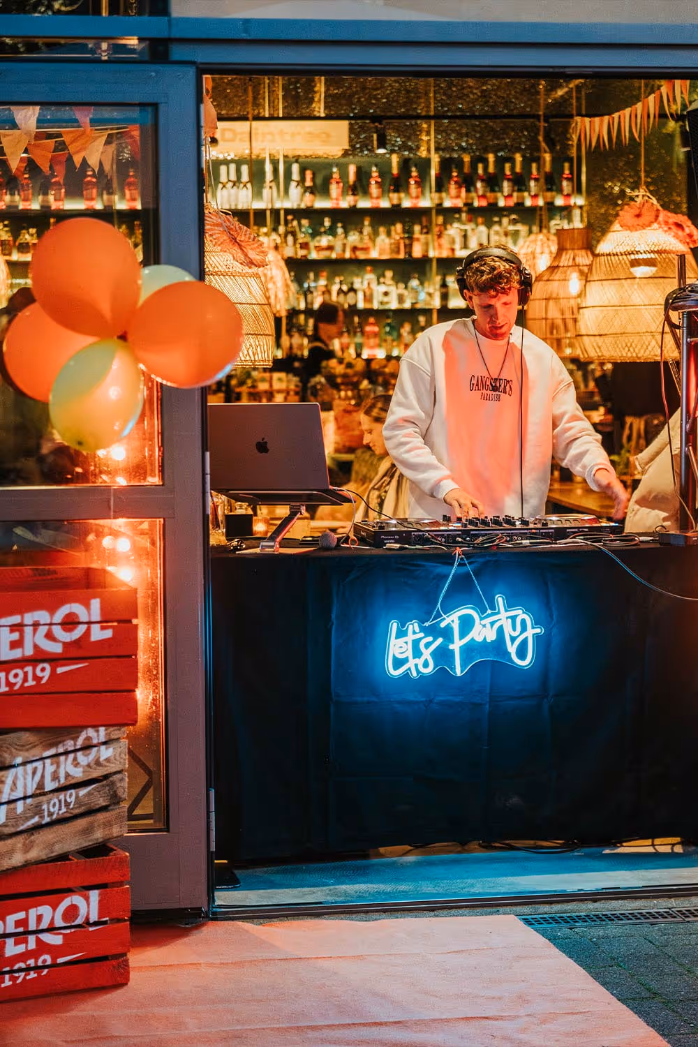 Ein DJ legt Musik auf einem Event auf, mit einem Laptop, Mischpult und leuchtendem Schild „Let's Party“ in einer Bar mit Regalen voller Flaschen.