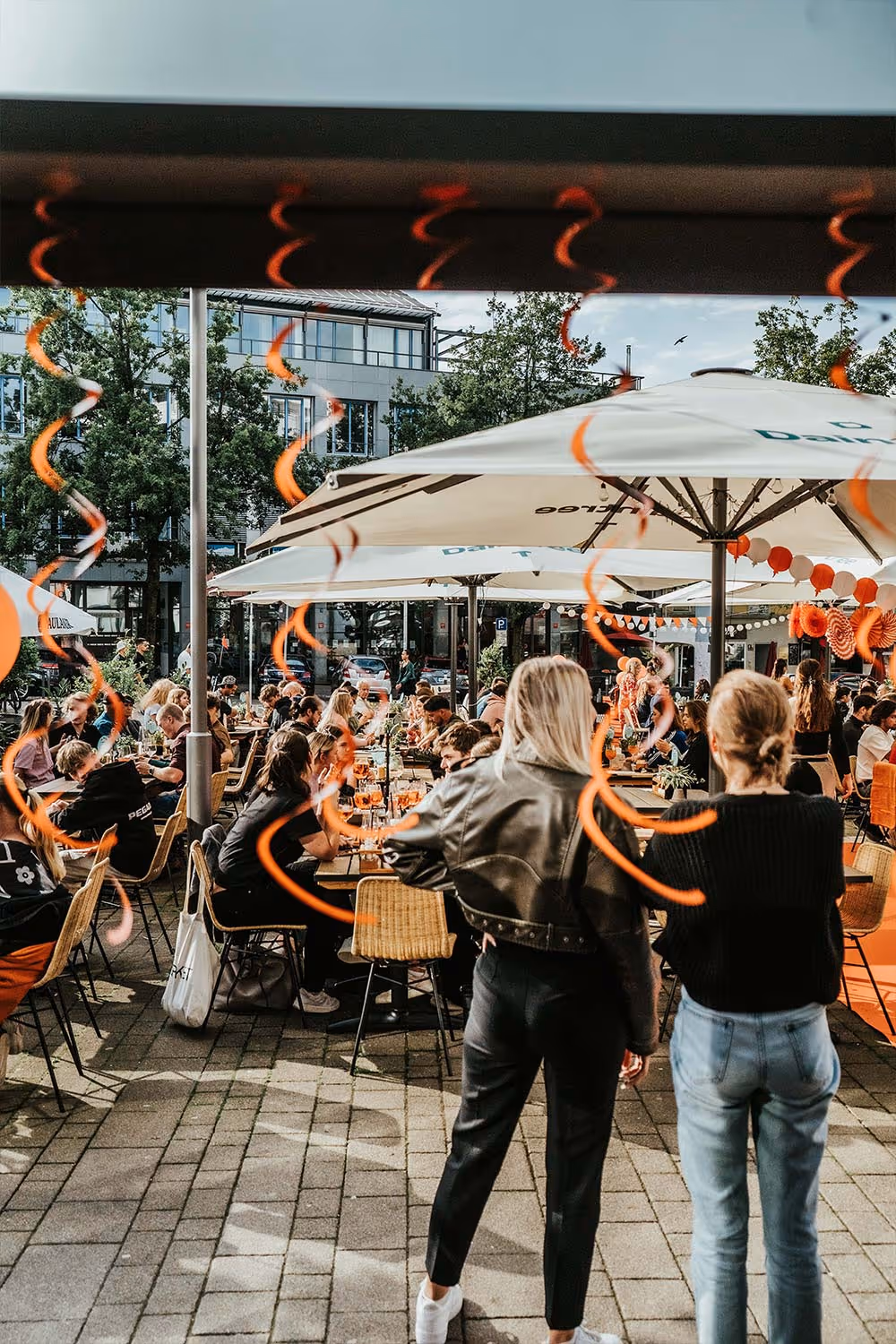 Menschen sitzen und unterhalten sich im Außenbereich eines Cafés mit Sonnenschirmen und orangener Dekoration.