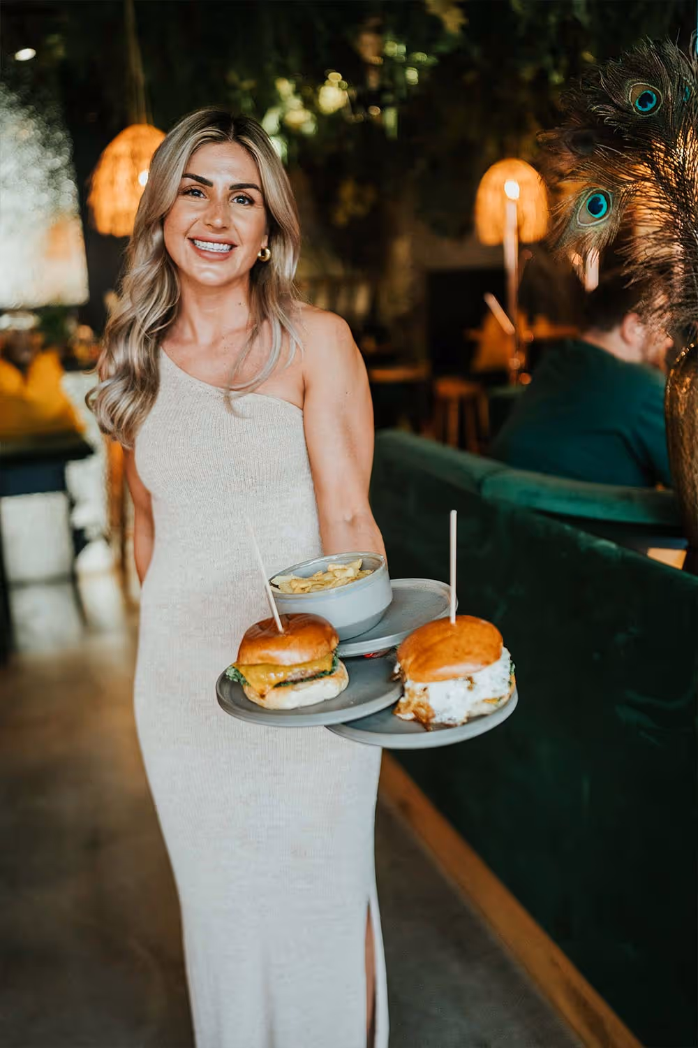 Freundliche Kellnerin in einem beigen, ärmellosen Kleid trägt zwei Burger und eine Schüssel Pommes in einem Restaurant.