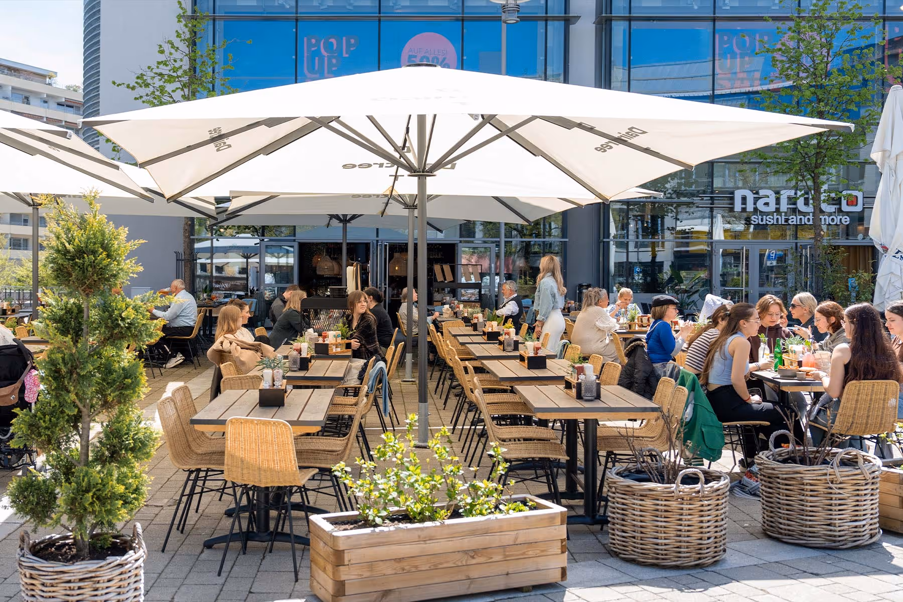 Menschen sitzen bei Sonnenschein an Holztischen unter großen weißen Sonnenschirmen im Außenbereich eines Restaurants namens Naruto Sushi and More.