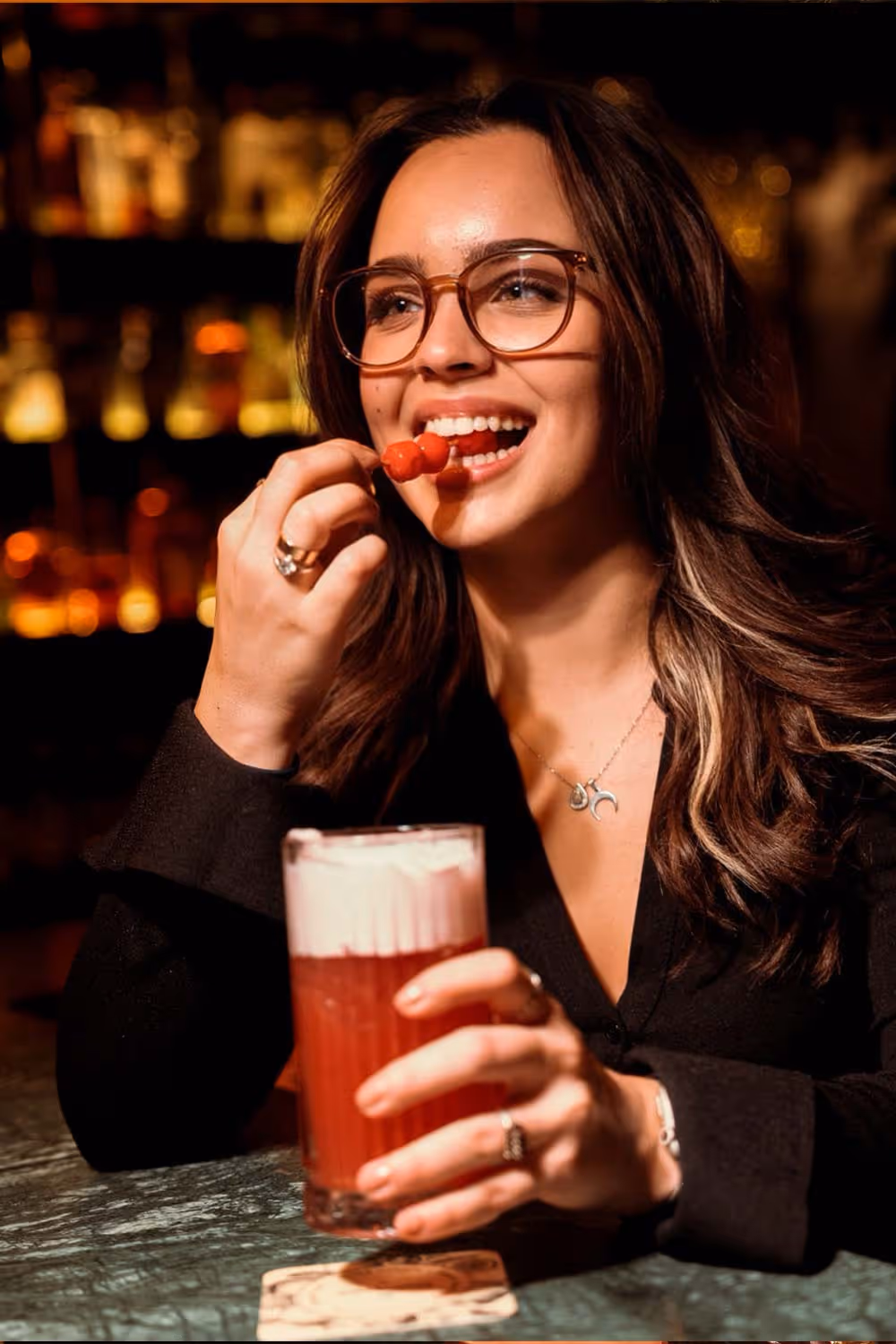 Frau mit Brille isst Cocktailkirschen und hält ein Glas mit rotem Getränk und Schaum in einer Bar.