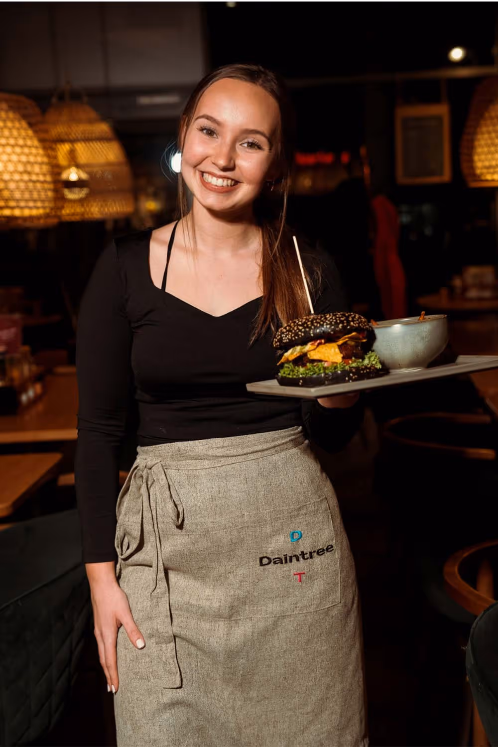 Lächelnde Kellnerin mit schwarzem Oberteil und grauer Schürze hält Tablett mit Burger und Schüssel in einem Restaurant.