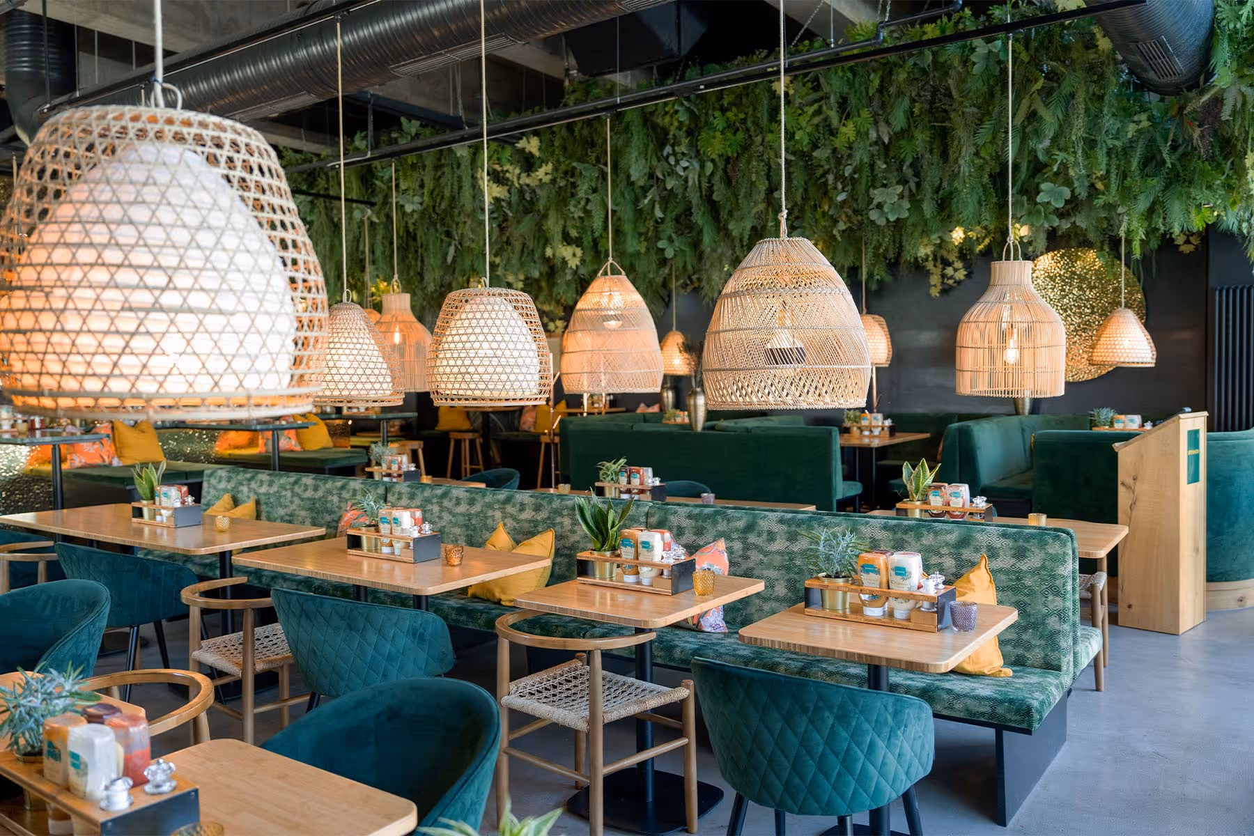 Modernes Café mit grünen Samtsesseln, Holztischen, hängenden Korblampen und einer Wand aus grünem Pflanzenlaub.