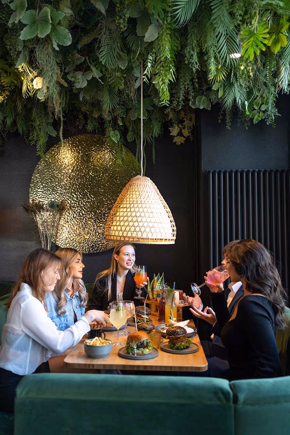 Fünf Personen sitzen an einem Tisch in einem Restaurant mit grünen Pflanzen an der Decke und essen Burger und Pommes, während sie Getränke genießen.