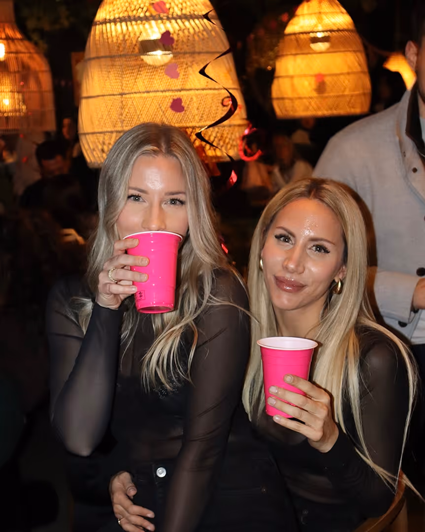 Zwei blonde Frauen in schwarzen, transparenten Oberteilen halten pinke Becher bei einer Party mit warmem Lampenlicht im Hintergrund.