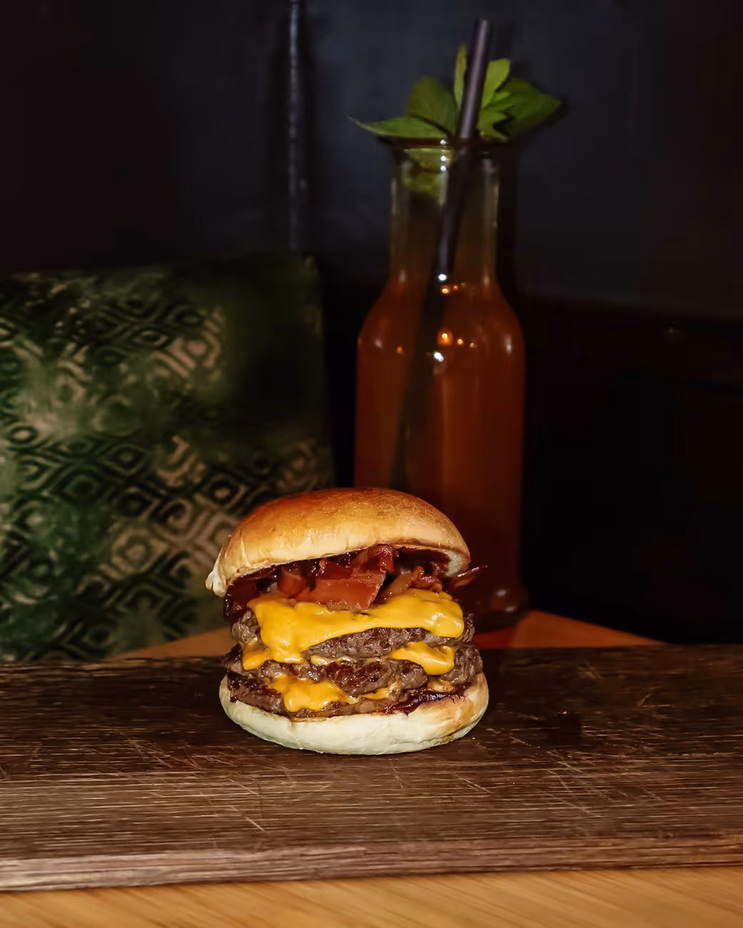 Doppelter Cheeseburger mit Speck auf einem Holzbrett, im Hintergrund ein Getränk mit Minzblättern.