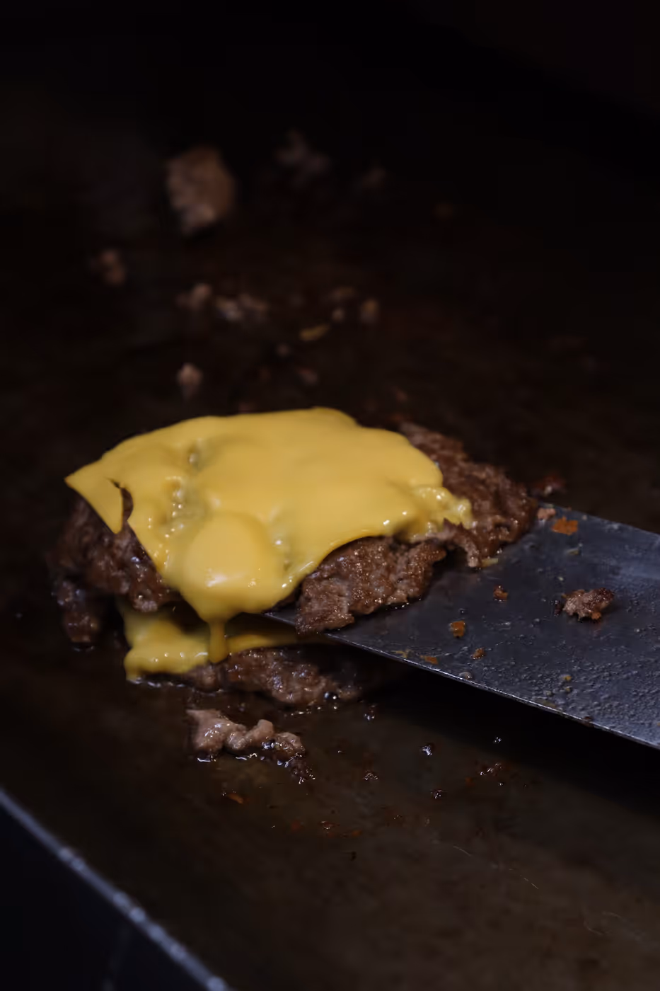 Doppelte Hamburger-Patties mit geschmolzenem Käse werden mit einem Metallspatel auf einer Grillplatte angehoben.