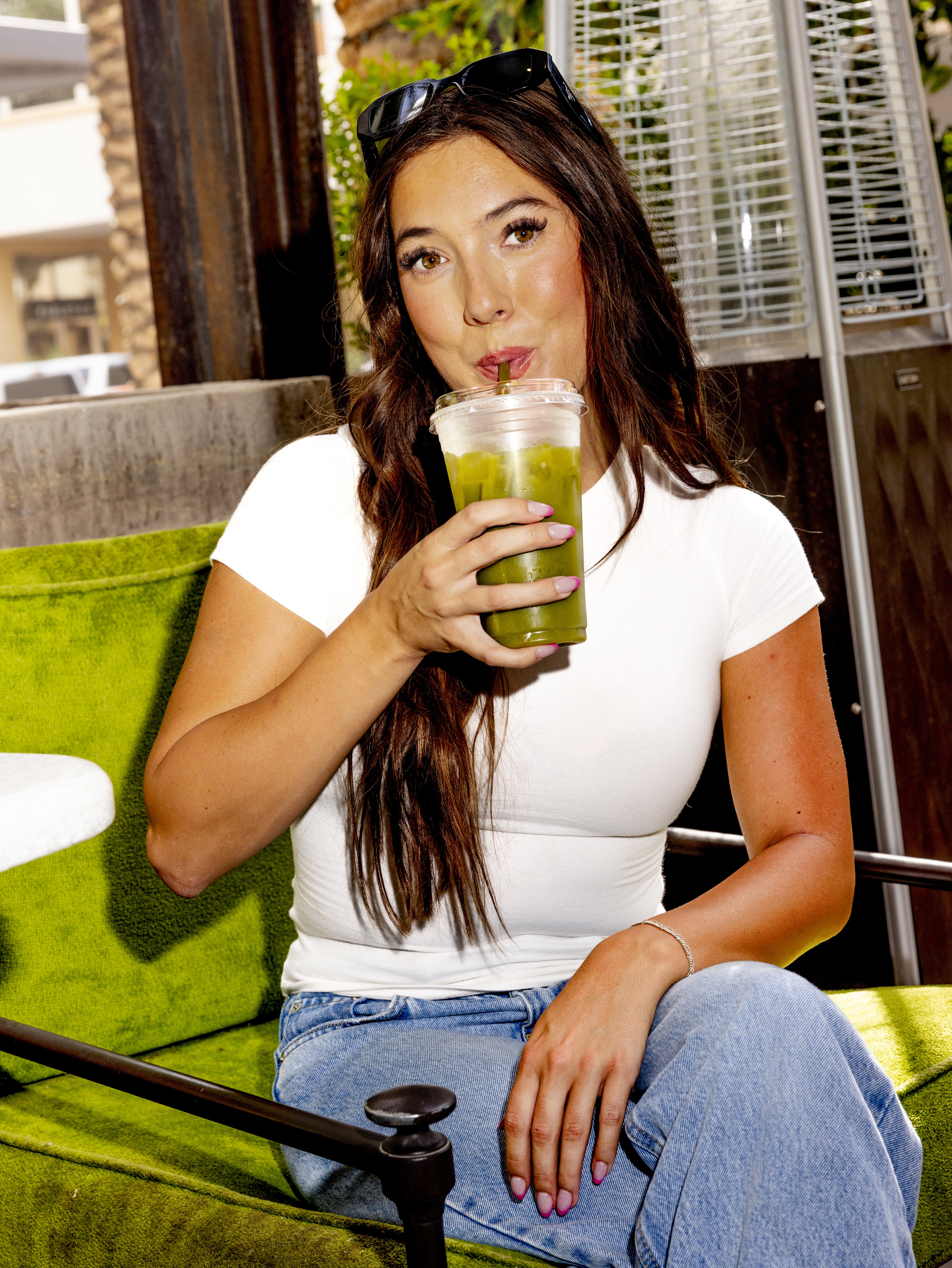 Woman with long brown hair sipping a green iced drink while seated on a green cushioned chair.