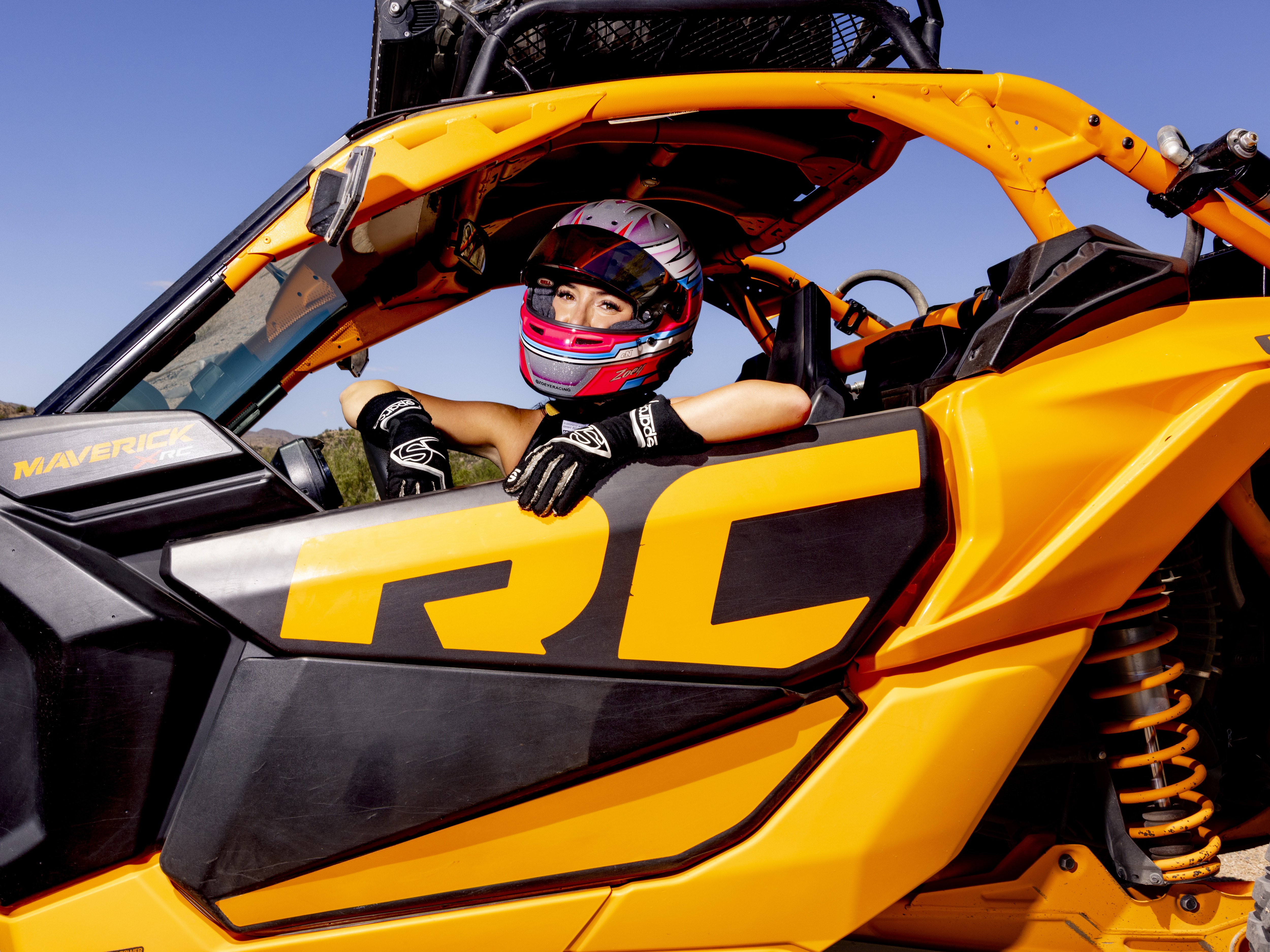 Person wearing a pink and white helmet and black gloves sitting inside an orange Maverick RC off-road vehicle.