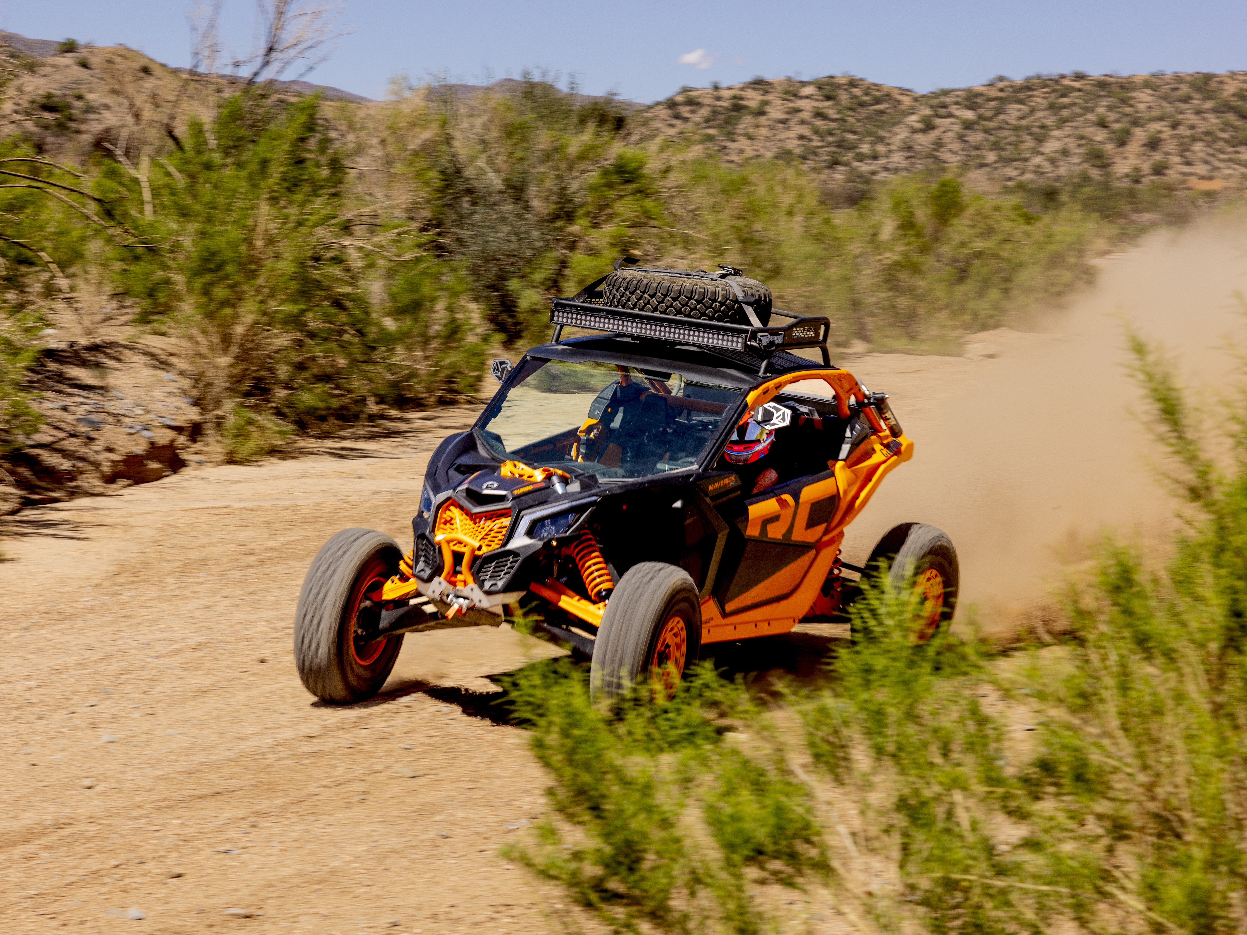 Orange and black off-road vehicle kicking up dust while racing on a dirt trail in a desert landscape.