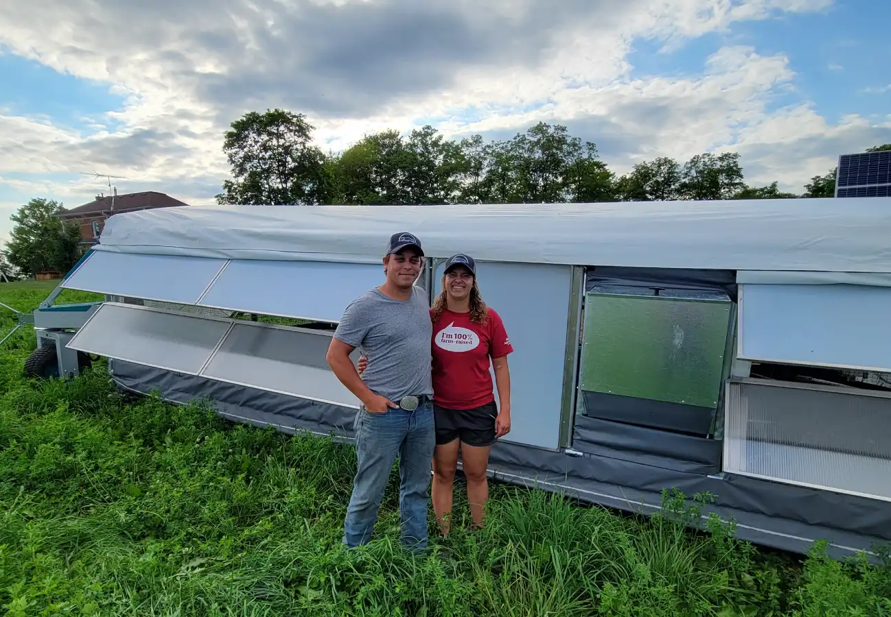 Casie and Tom De Boer standing together in front of their ROVA|BARN on a green pasture under a partly cloudy sky.