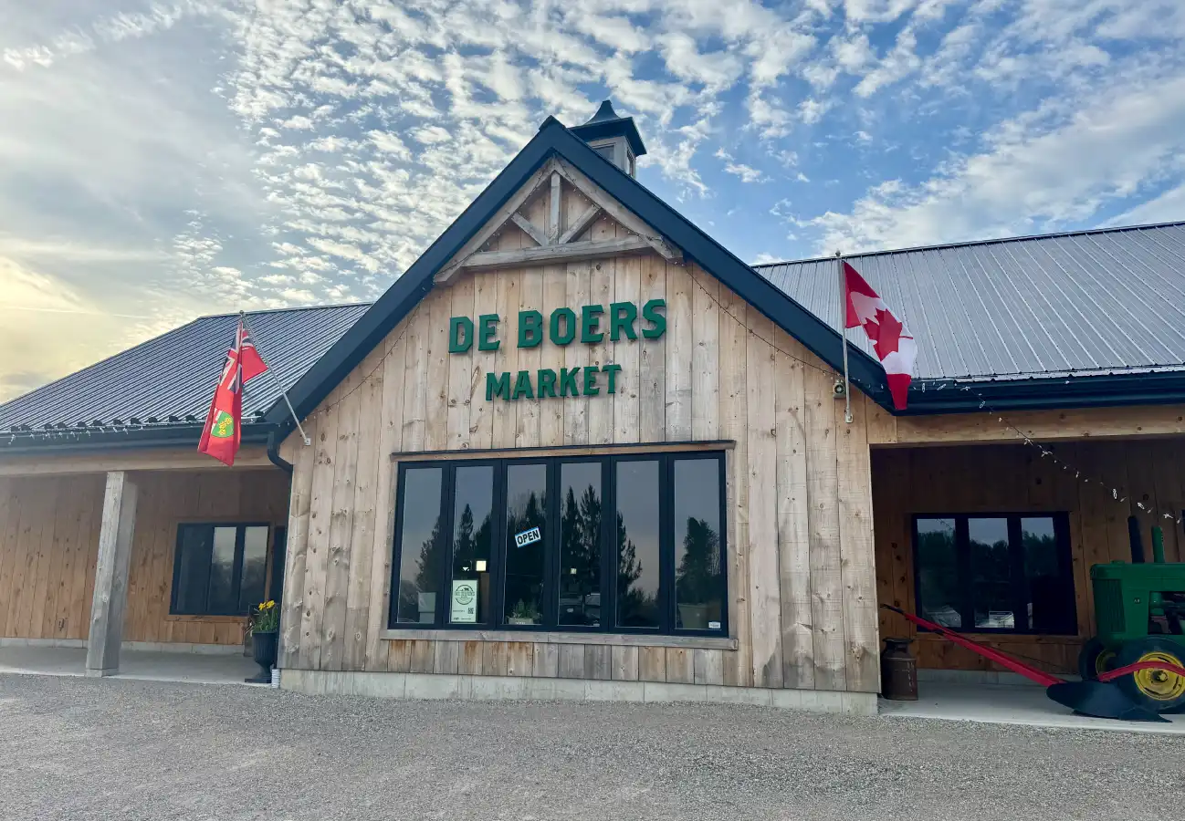 Exterior of the De Boers Market storefront, featuring a wood facade, Canadian and Ontario flags, and a vintage green tractor under a blue sky.
