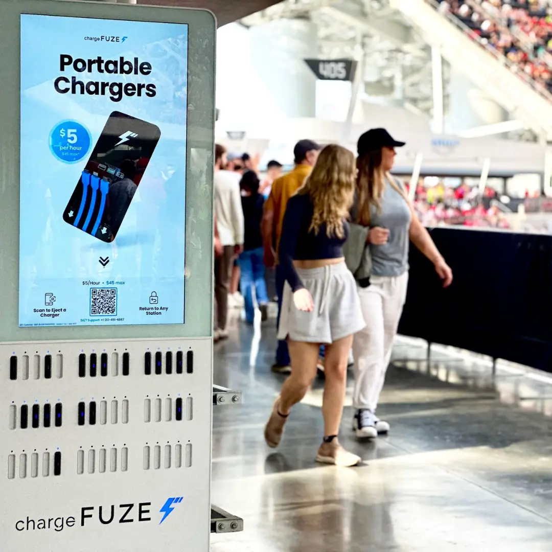 Cell phone charging station kiosk inside stadium concourse with fans walking past in the background.