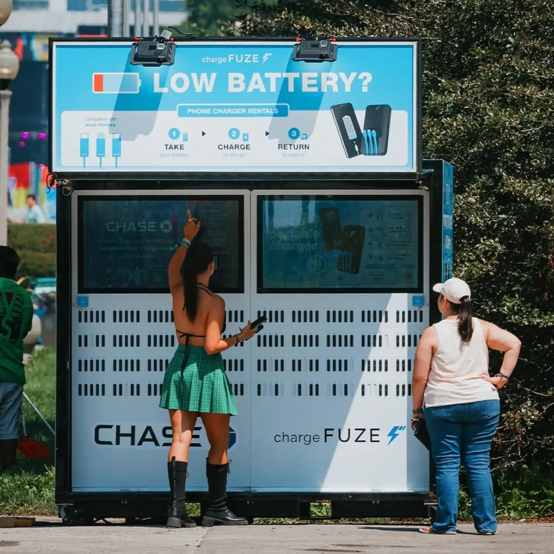 Phone charging stations outdoors at a festival with attendees renting portable chargers from a large kiosk.