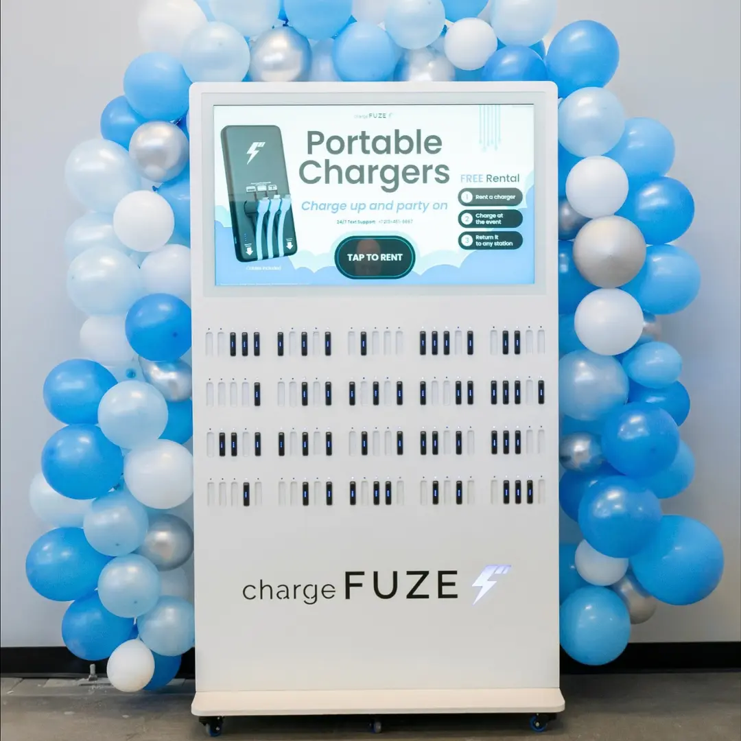 Phone charging station by ChargeFUZE offering free portable charger rentals displayed in front of a blue and silver balloon arch at an event venue