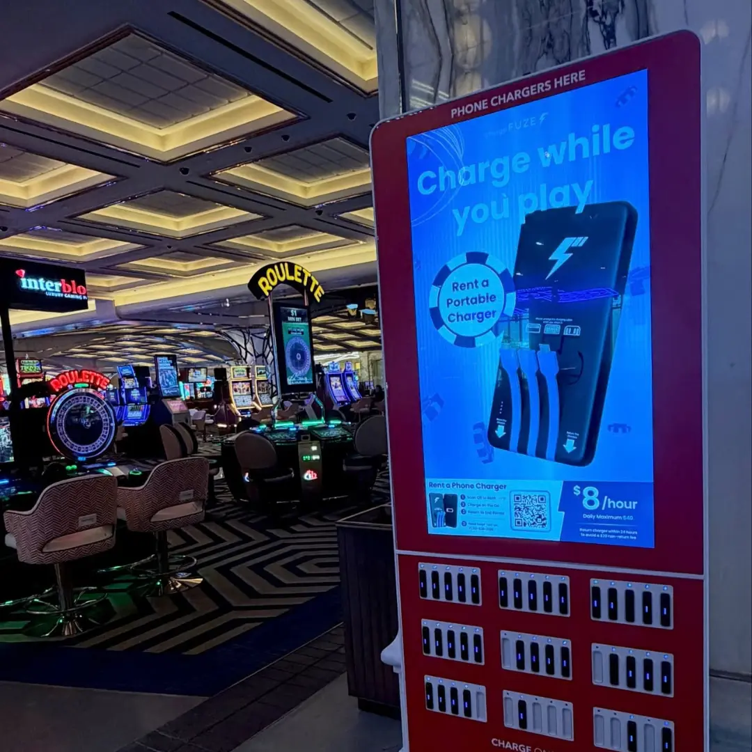 Cell phone charging station kiosk by ChargeFUZE offering portable charger rentals for $8 per hour at the entrance of a casino floor with roulette tables visible in the background 