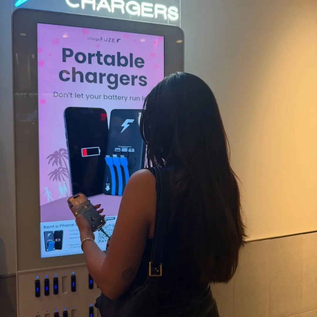 Phone charging station kiosk by ChargeFUZE as a woman holds her smartphone while browsing the portable charger rental screen