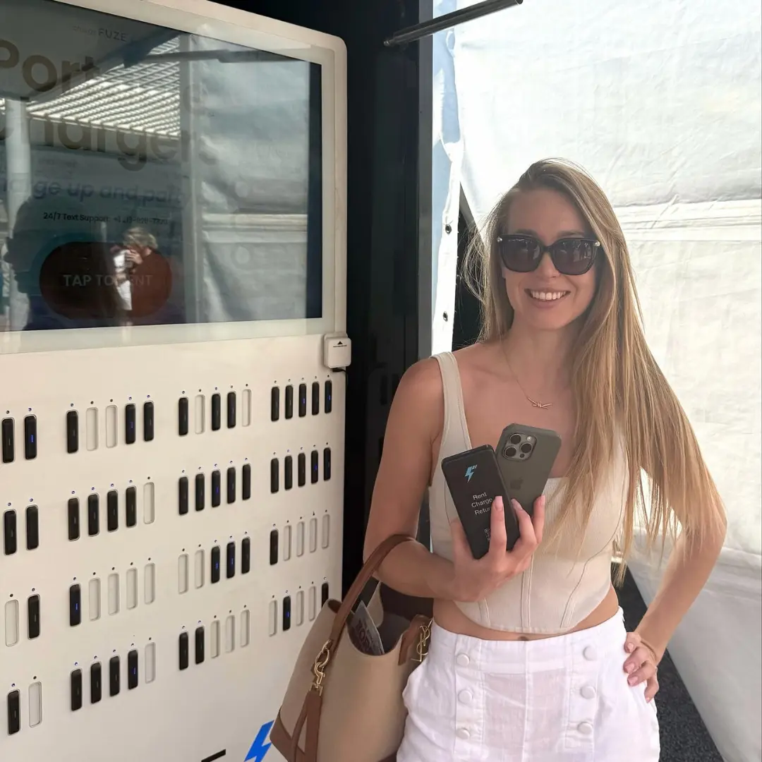 Smiling woman holding a rented ChargeFuze portable charger next to a mobile power station kiosk at an outdoor event.