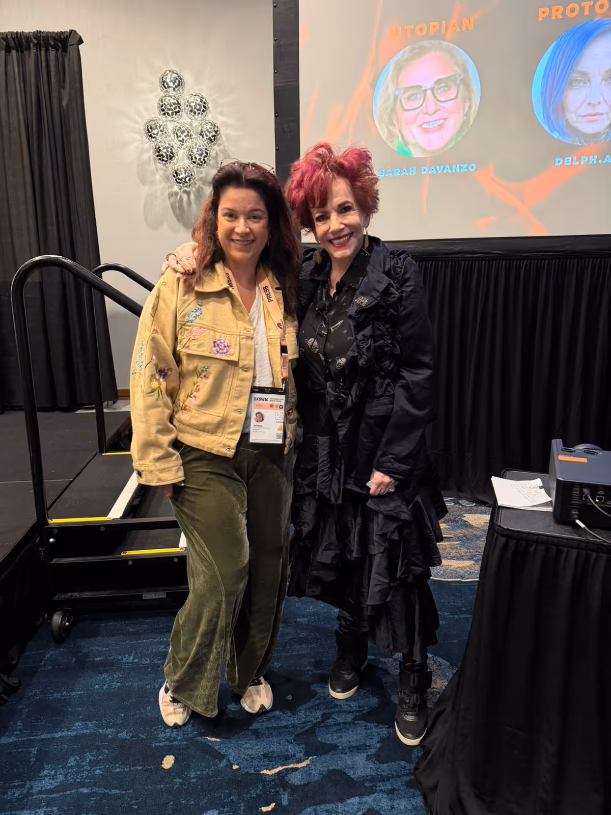 Duas mulheres posando juntas no SXSW 2026, uma com jaqueta bege bordada e outra com vestido preto e cabelo vermelho.