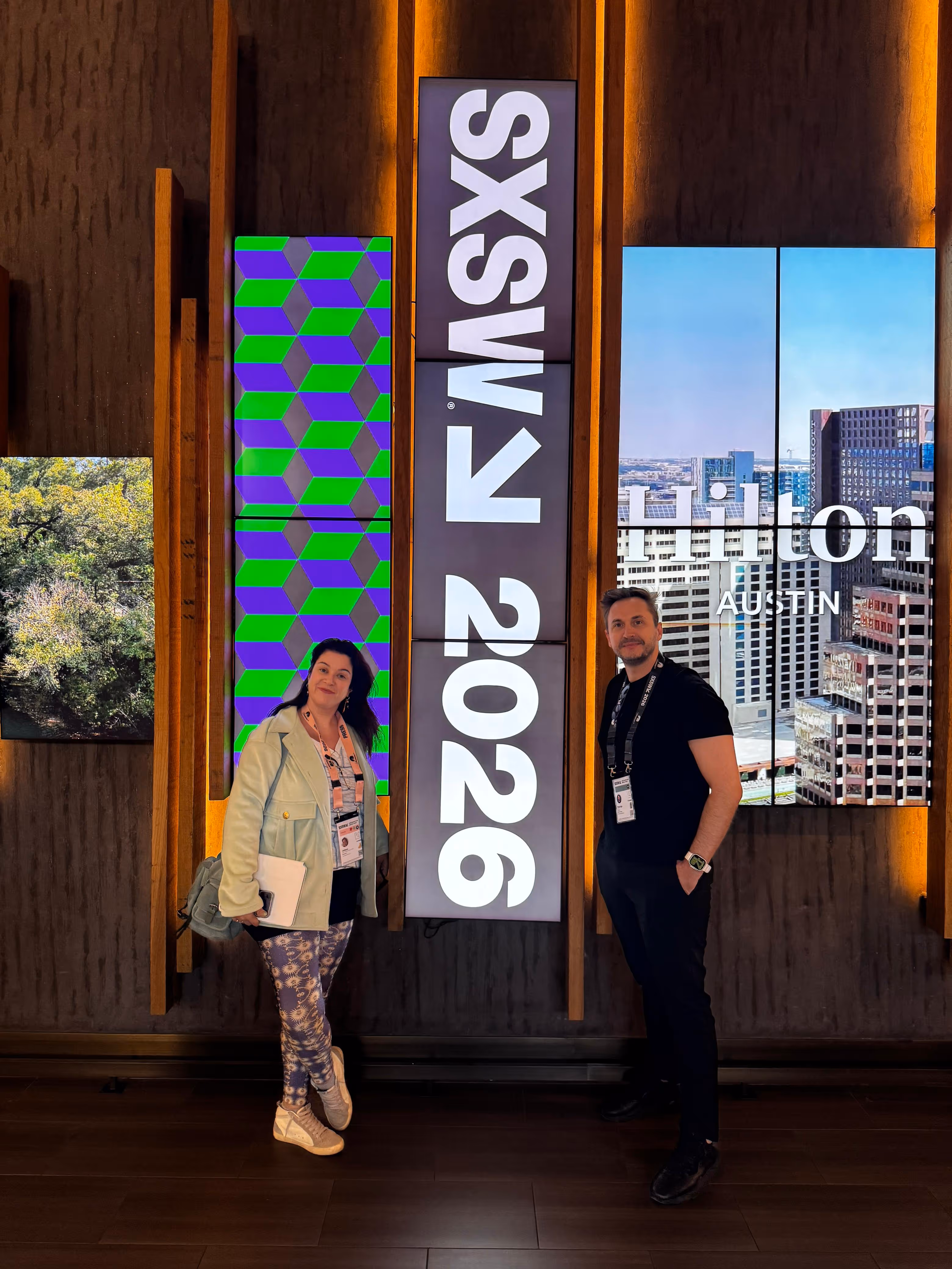 Duas pessoas em pé em frente a painéis digitais com texto 'SXSW 2026'