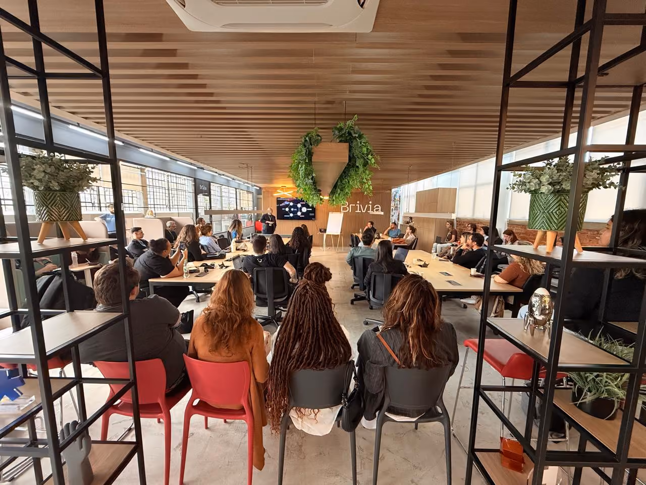 Sala da Brivia no Instituto Caldeira durante a Semana Caldeira 2025. Sala moderna com teto de madeira, plantas penduradas no centro e várias pessoas sentadas em cadeiras ao redor de uma mesa, assistindo a uma apresentação em uma tela na parede.