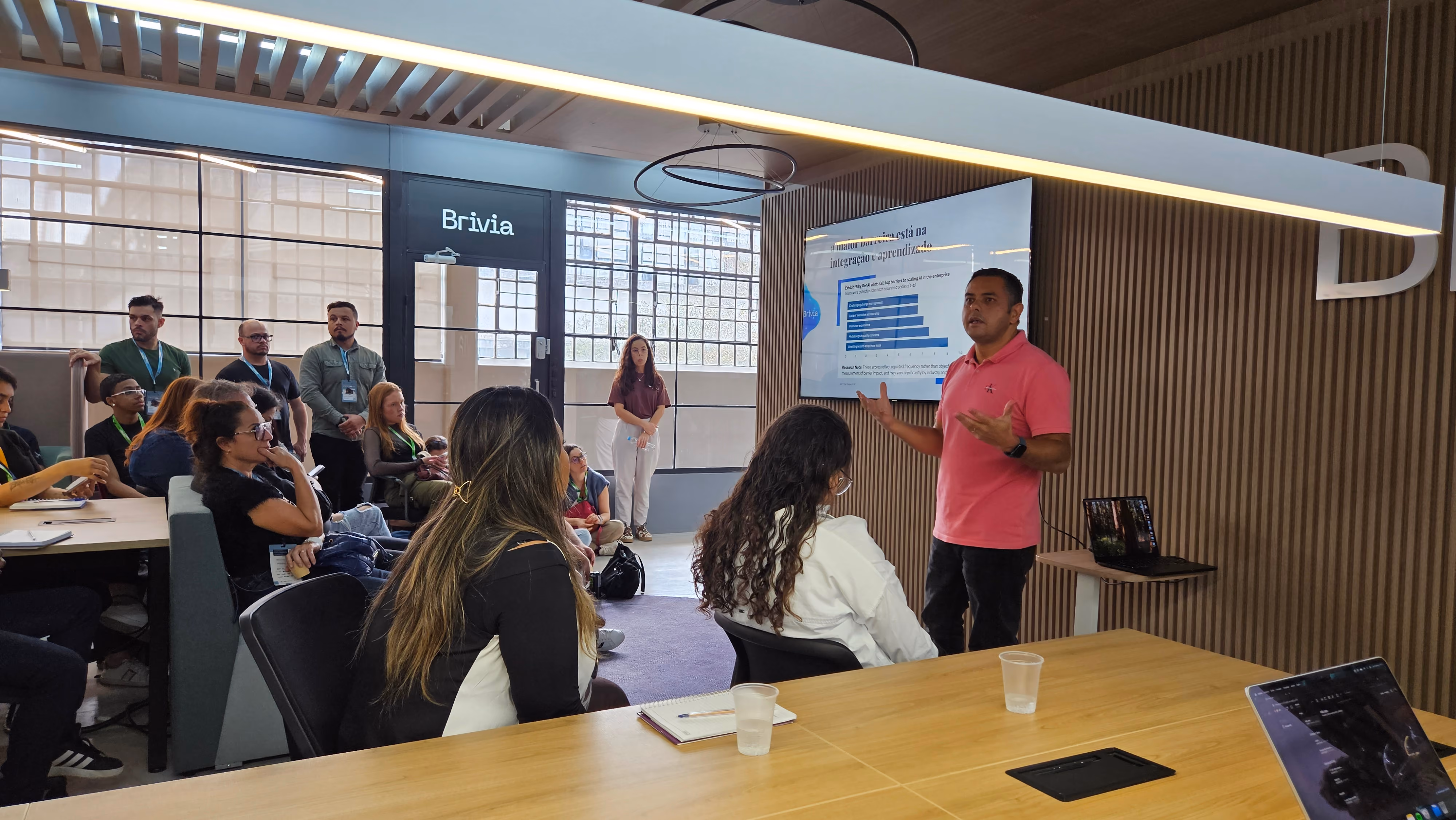Roberto Ribas apresenta dados em uma tela para grupo sentado na sala da Brivia, com paredes de madeira e grande janela ao fundo.