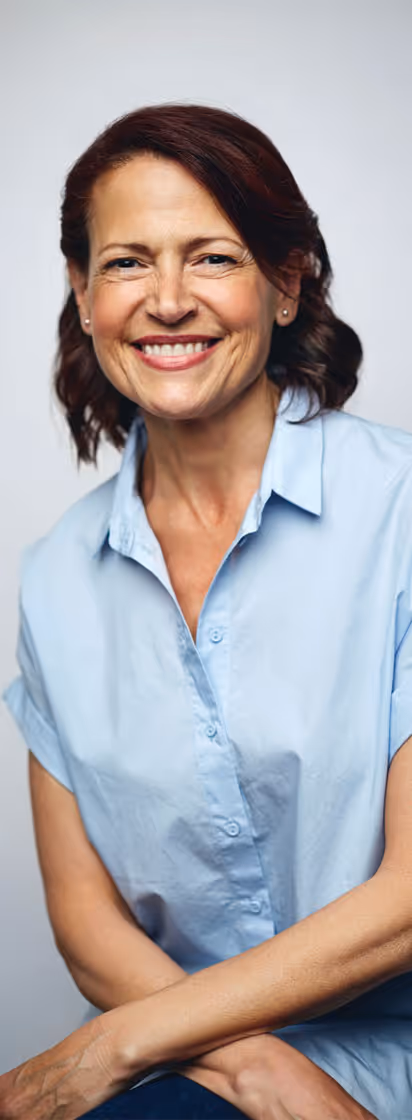 A middle-aged woman with auburn hair smiling warmly in a light blue shirt, posed casually against a light background.