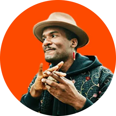 Man wearing a beige fedora and patterned jacket smiling with hands clasped against an orange circular background.