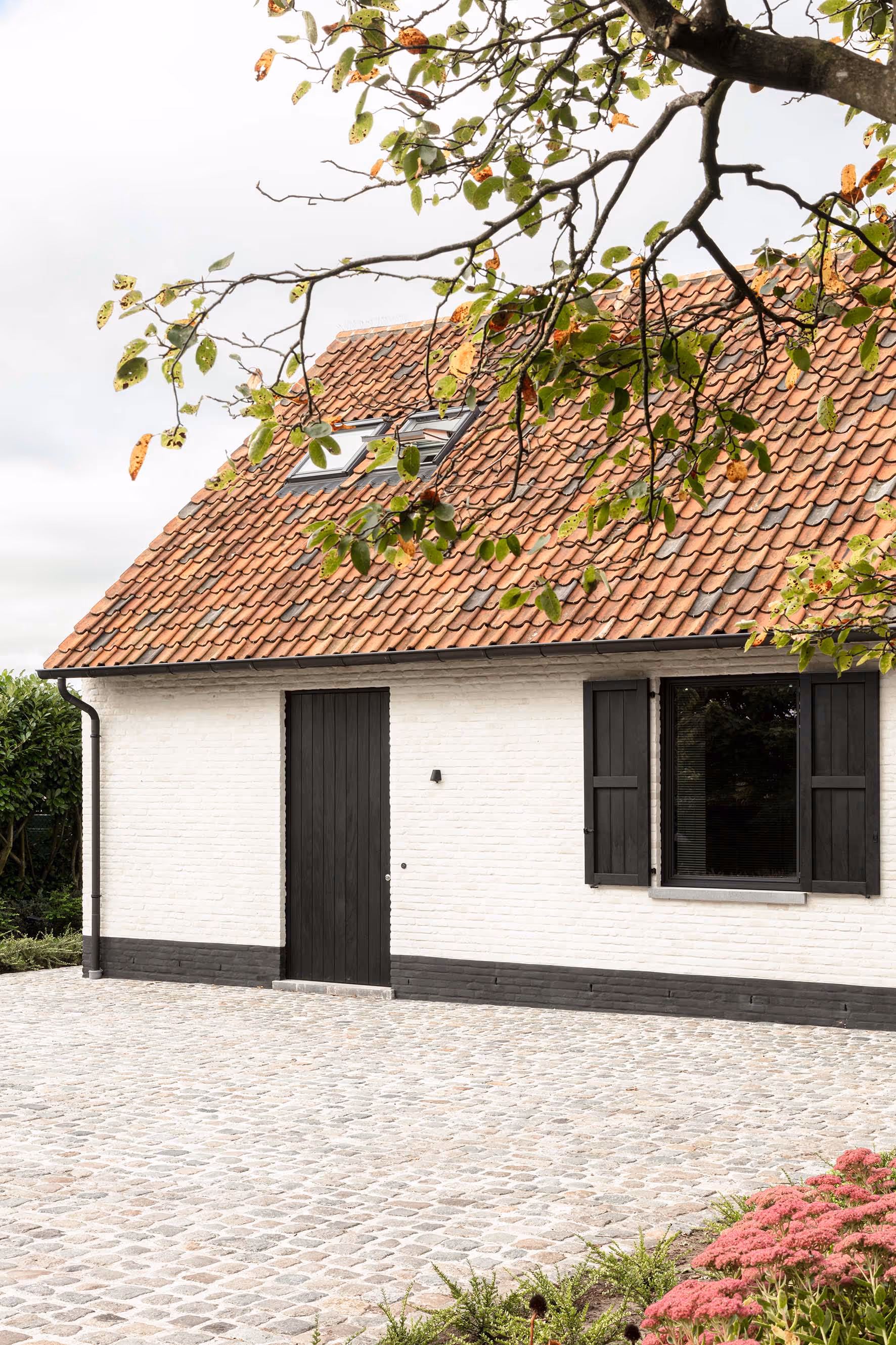 Traditioneel wit huis met zwarte luiken, kasseien oprit en herfstbladeren.